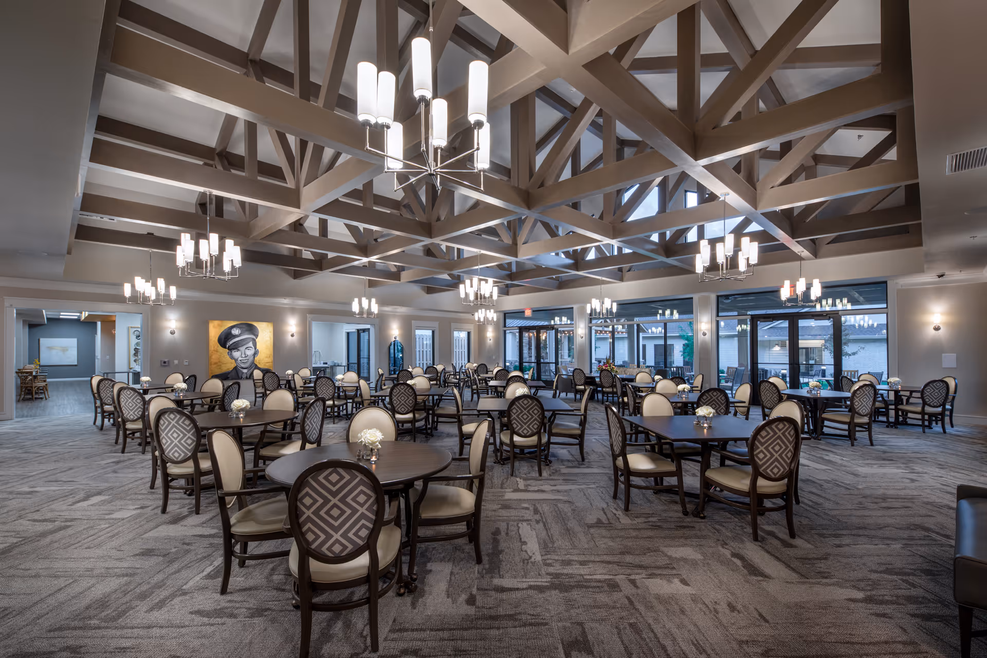 Spacious dining room with multiple round tables and cushioned chairs arranged neatly. The room features a high ceiling with exposed wooden beams and modern chandeliers hanging down. Large windows and glass doors allow natural light to enter, and there is a portrait on one wall. The carpeted floor and neutral color palette create a warm and inviting atmosphere.
