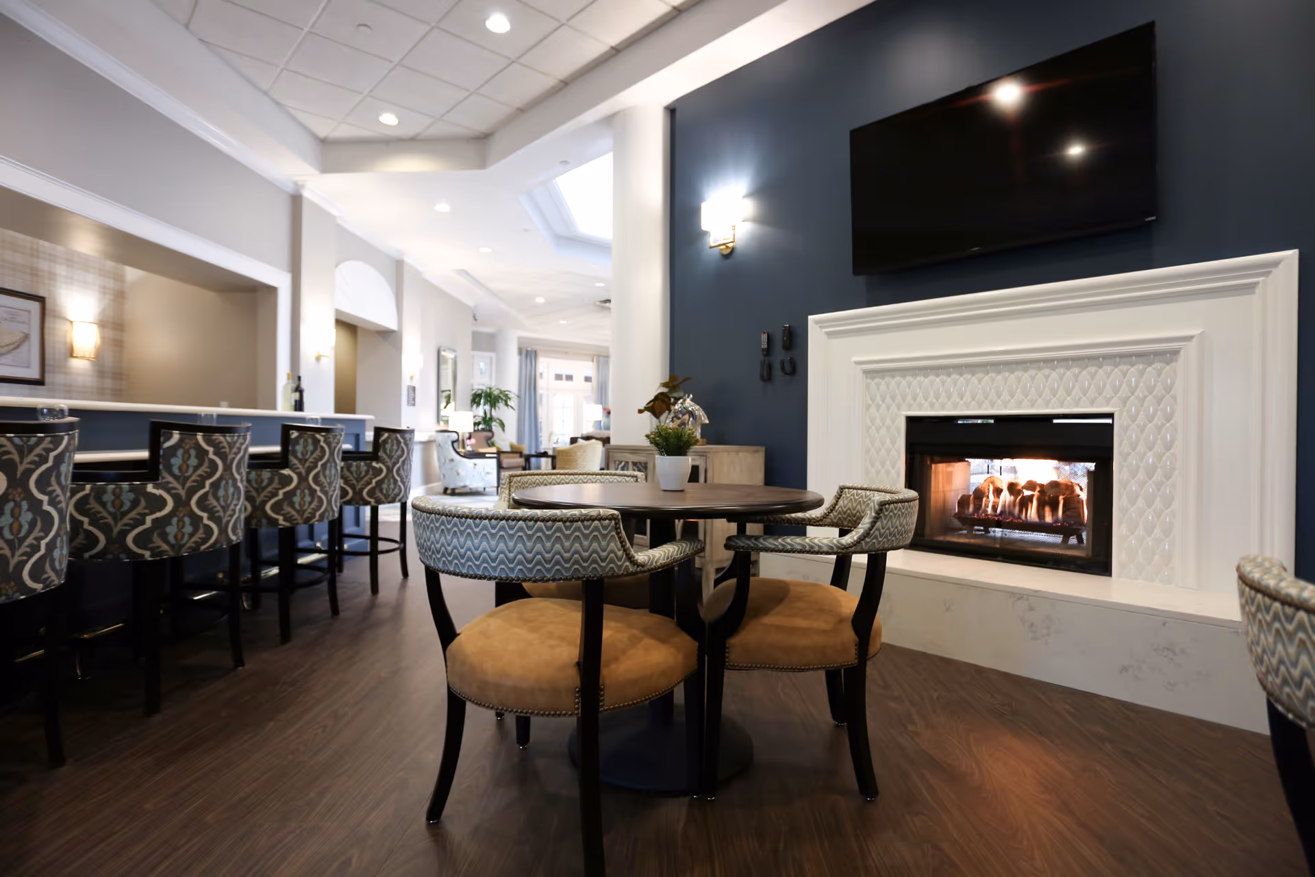 A cozy interior common area with a round table surrounded by four cushioned chairs, a modern fireplace with a white decorative mantel, and a flat-screen TV mounted above it. To the left, there is a bar counter with patterned bar stools and soft lighting, with a bright hallway extending into the background.