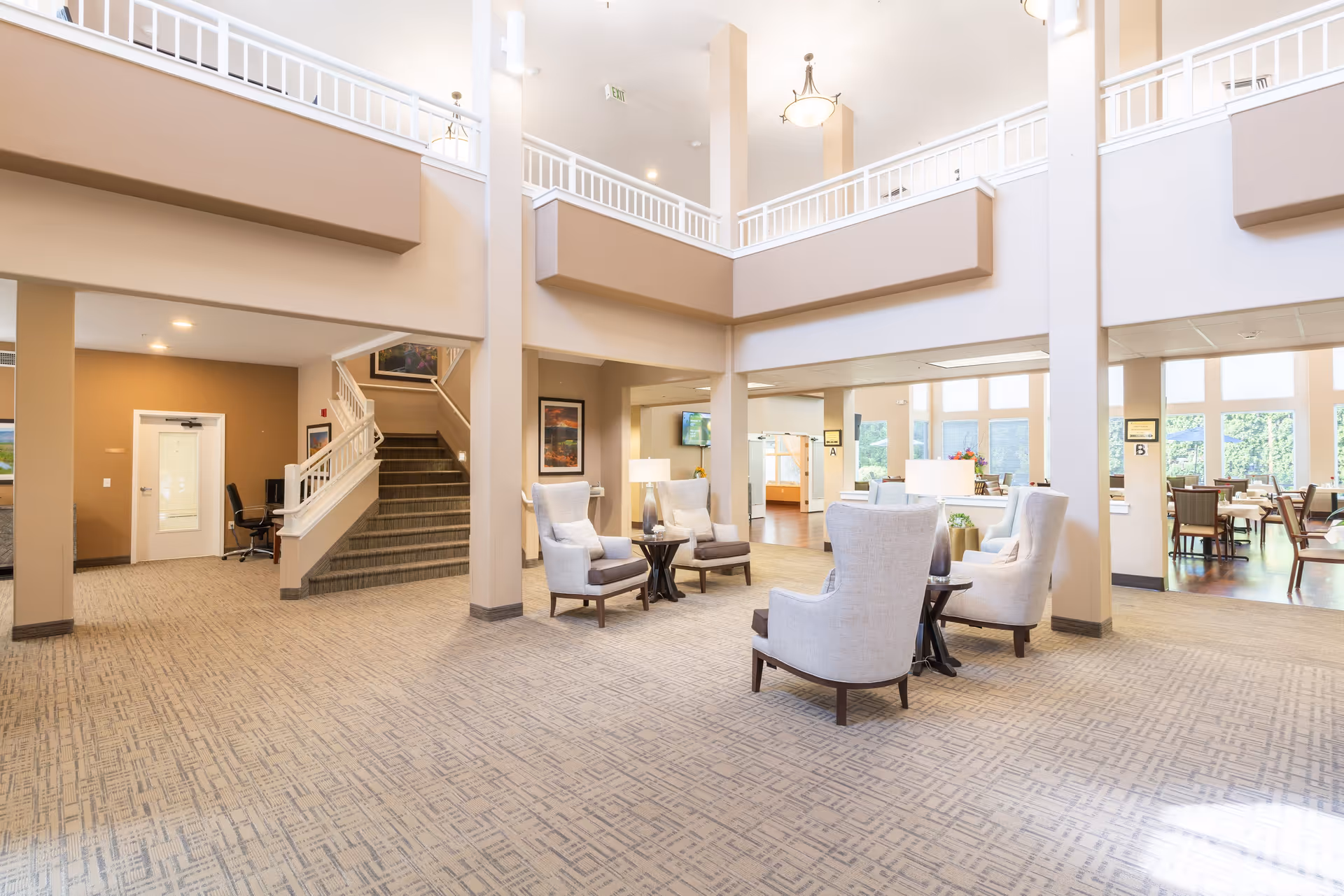 Bright and spacious senior living facility common area with high ceilings, beige walls, and carpeted floors. The area features several comfortable armchairs arranged around small tables with lamps. There is a staircase leading to an upper floor, large windows letting in natural light, and a dining area visible in the background.