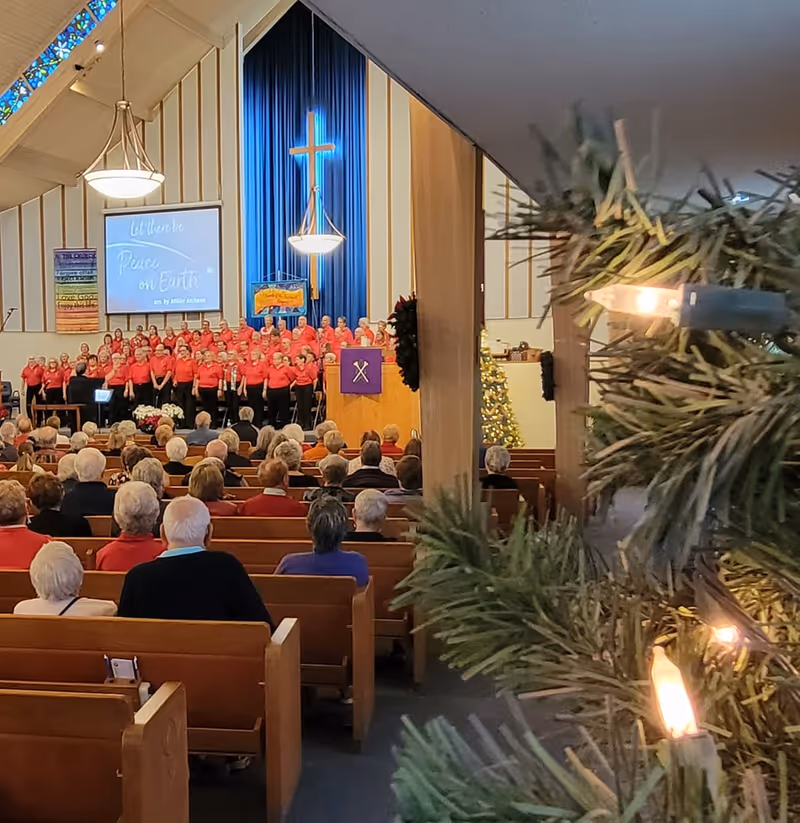 Interior of a church with a choir dressed in red shirts standing on the stage in front of a large illuminated cross and blue curtains. The congregation, mostly elderly people, is seated in wooden pews facing the choir. A Christmas tree with lights is partially visible on the right side of the image.