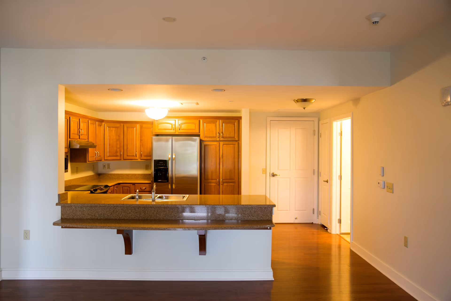 Interior view of a senior living community kitchen with wooden cabinets, a stainless steel refrigerator, a double sink on a granite countertop, and hardwood floors. There are two doors visible in the background and warm lighting illuminating the space.