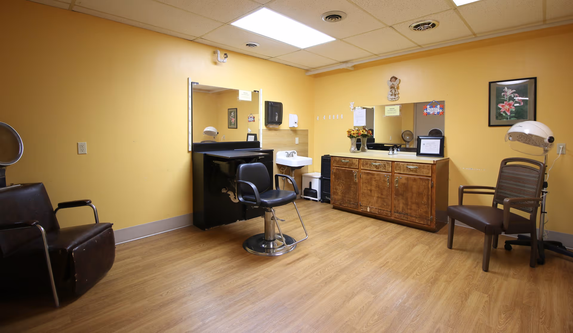 Interior of a hair salon room with yellow walls and wood flooring. The room contains a black salon chair in front of a mirror and sink, a wooden cabinet with a mirror above it, two additional chairs, a hair dryer, and some wall decorations including a framed flower picture.