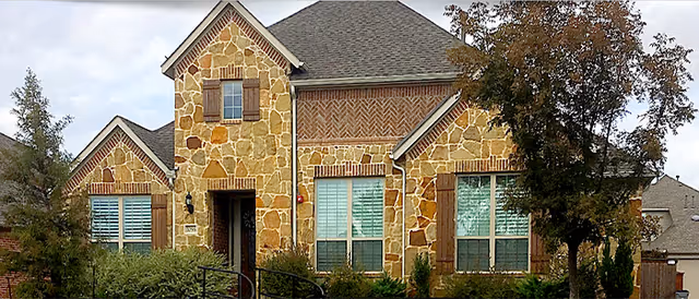 Stone-faced residential building with stone accents, windows, trees, and landscaping in front.