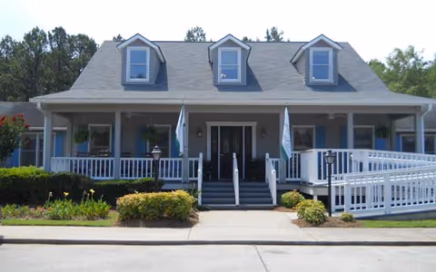 Front exterior view of a two-story building with a gray roof and blue siding. The building has three dormer windows on the roof and a covered porch with white railings and steps leading up to double front doors. There are two flags on poles on either side of the steps and some landscaping with bushes and flowers in front.