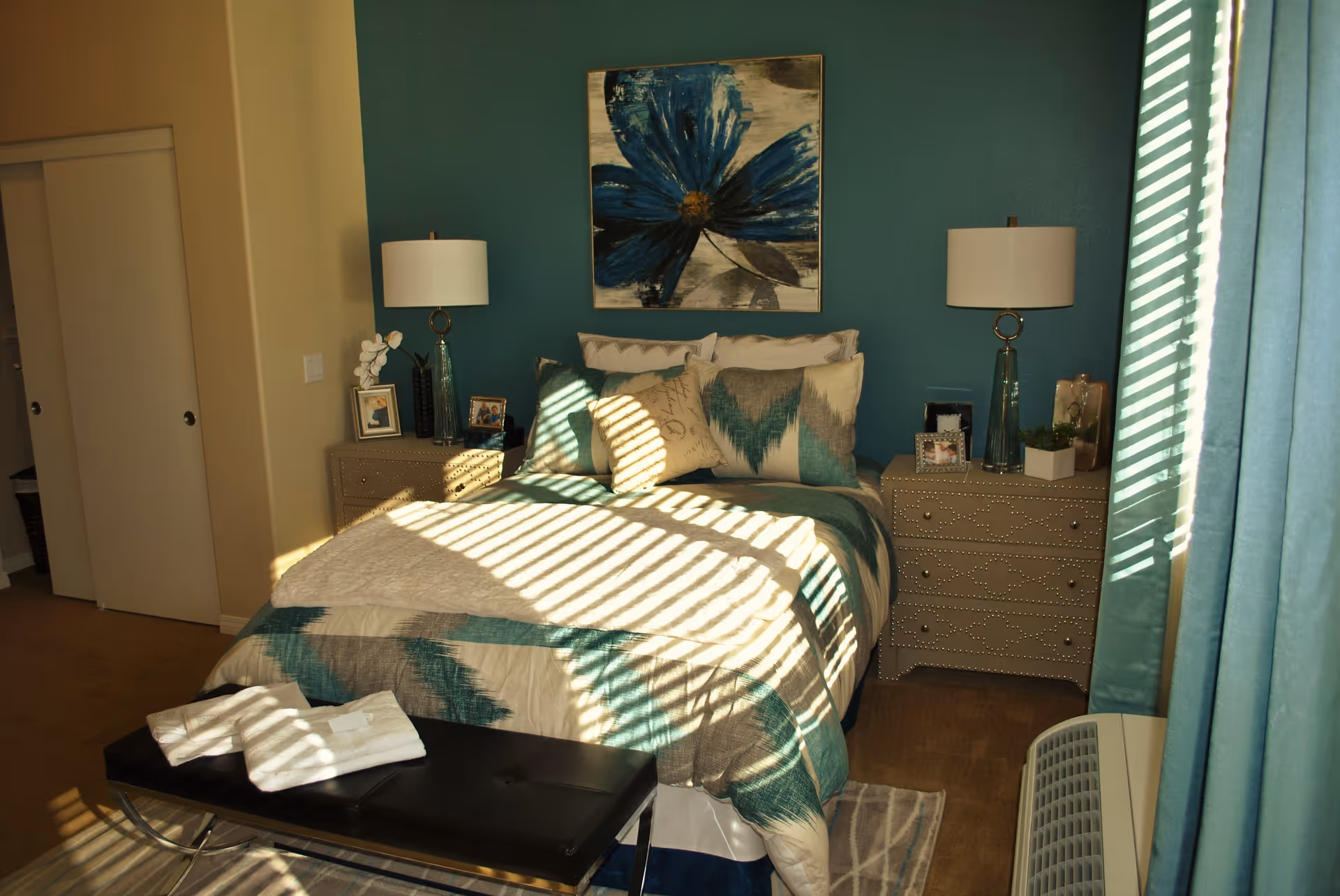 A cozy bedroom with a bed covered in a blue and beige patterned comforter and multiple pillows. There are two matching nightstands on either side of the bed, each with a lamp and decorative items. A large blue flower painting hangs on the teal accent wall behind the bed. Sunlight streams through window blinds, casting striped shadows across the bed and floor. A black bench with folded towels sits at the foot of the bed, and teal curtains frame the window on the right side.