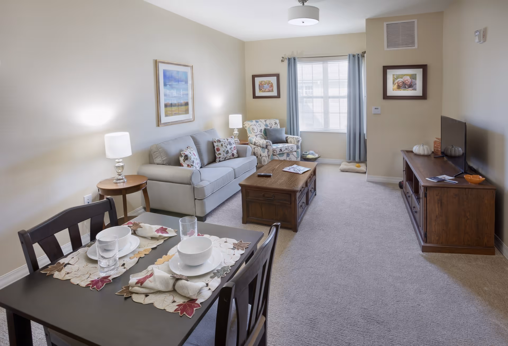 A cozy living room and dining area in an assisted living facility. The room features a gray sofa with floral pillows, a patterned armchair, a wooden coffee table, and a wooden TV stand with a flat-screen TV. A dining table with two place settings, including bowls, plates, glasses, and napkins, is in the foreground. The walls are light beige with framed pictures, and a window with blue curtains lets in natural light. The carpet is light gray, and there are two table lamps providing additional lighting.