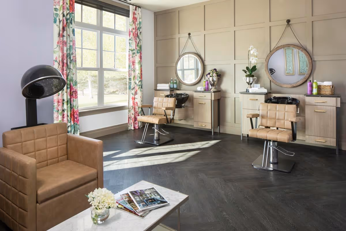 A bright and modern hair salon area with two styling chairs in front of round mirrors mounted on a beige paneled wall. Each station has a black sink and wooden cabinetry with hair products and towels. A large window with floral curtains lets in natural light. A brown leather armchair and a small table with magazines and a flower vase are in the foreground.