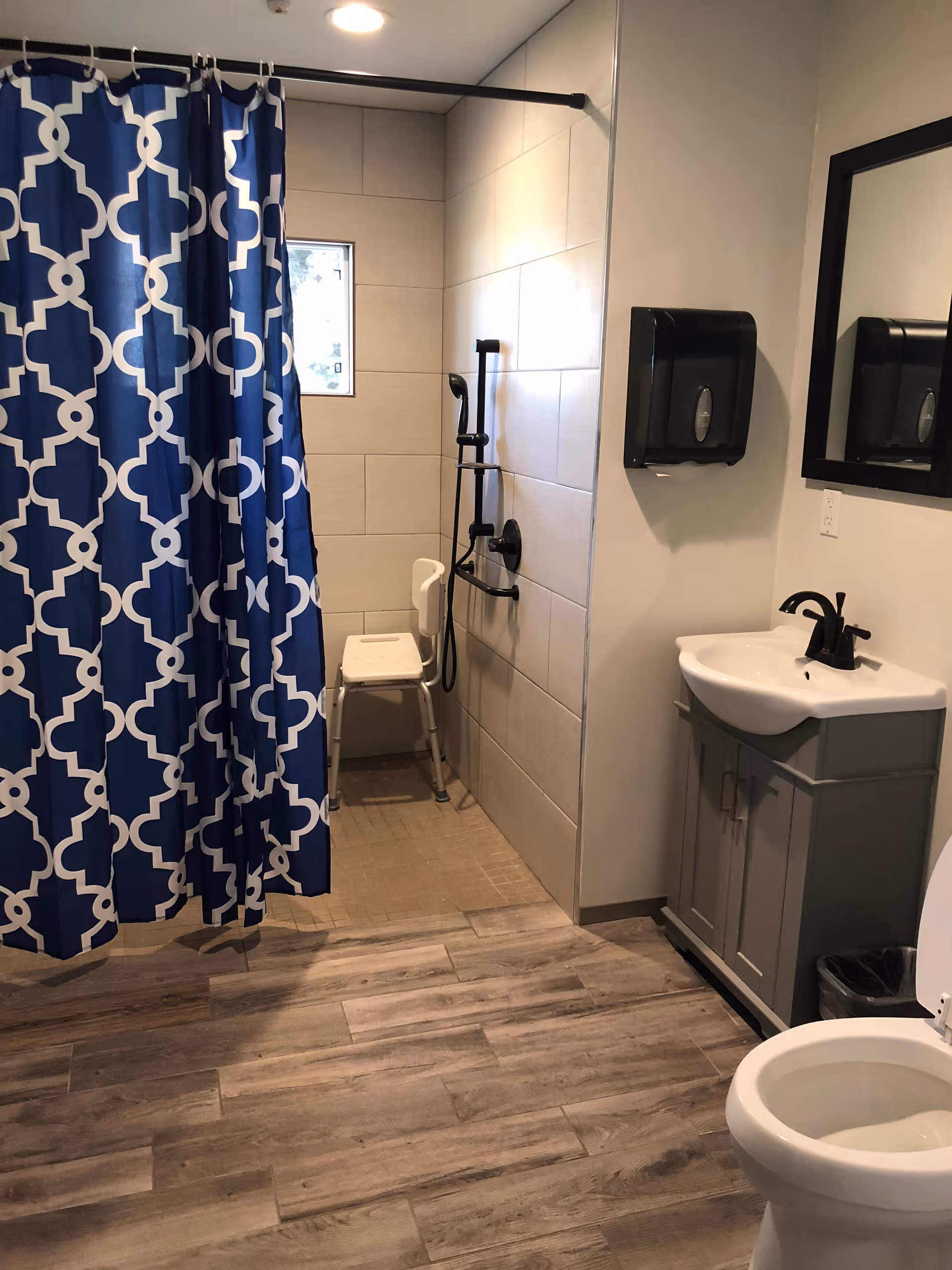A bathroom with a walk-in shower featuring a blue and white patterned shower curtain, a white shower chair, and a handheld showerhead. The bathroom also includes a white toilet, a gray vanity with a white sink, a black faucet, a black-framed mirror, and a black paper towel dispenser mounted on the wall. The floor has wood-like tiles and the walls are light-colored.