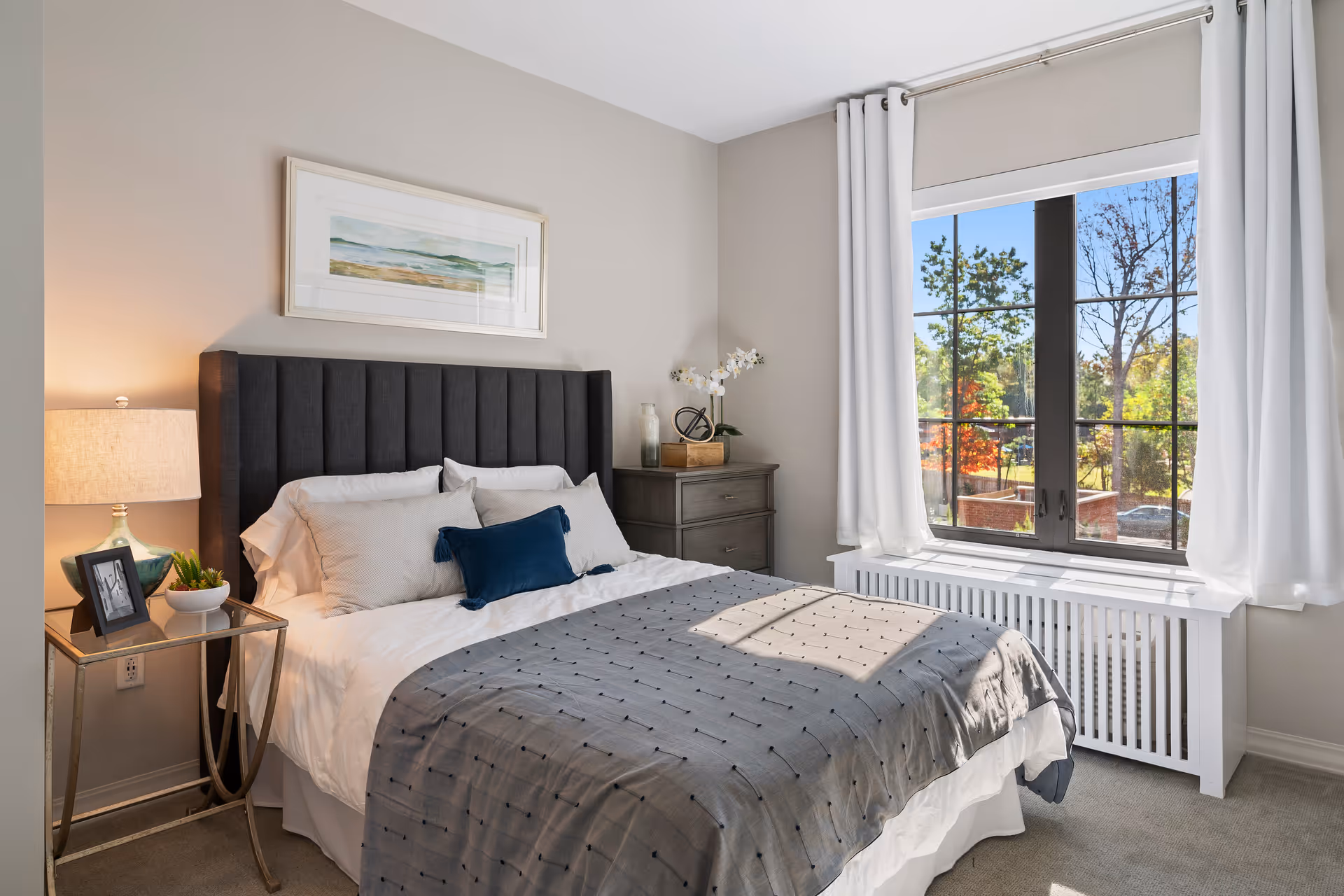 A bright and cozy bedroom with a large window showing a view of trees outside. The room features a bed with a dark upholstered headboard, white and gray bedding with a blue accent pillow, a nightstand with a lamp, a small plant, and a framed photo. There is a dresser with decorative items and white curtains framing the window.