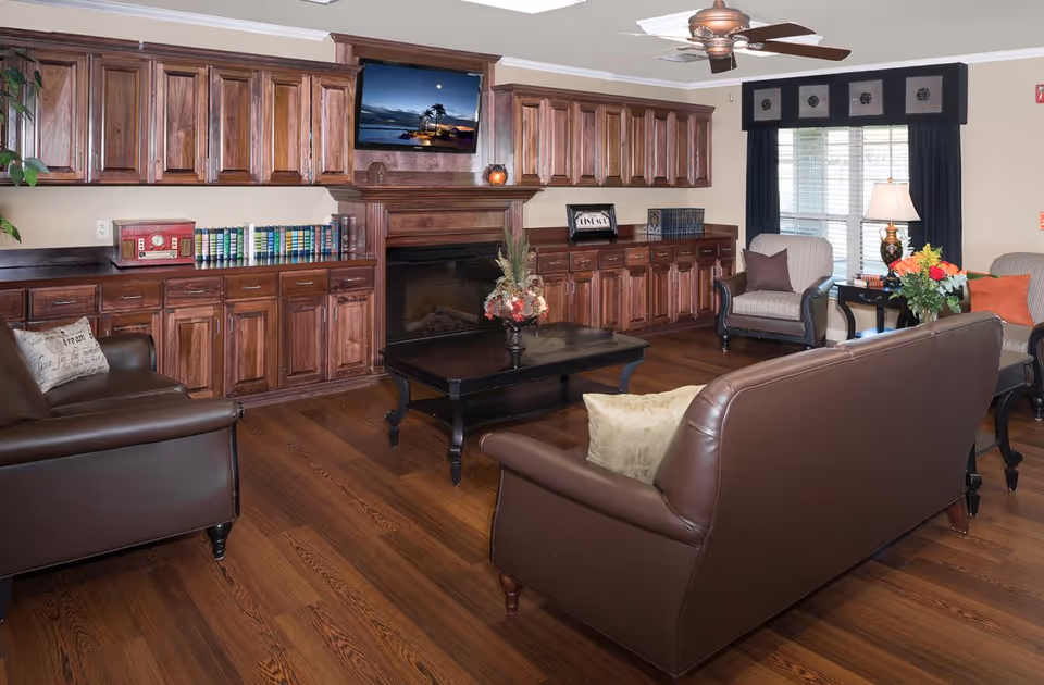 A cozy living room with wooden flooring and dark brown leather sofas arranged around a black coffee table with a floral centerpiece. The room features a wooden fireplace with a mounted TV above it, flanked by extensive wooden cabinetry filled with books and decorative items. There are two armchairs near a window with dark curtains, a side table with a lamp, and a vase of flowers.