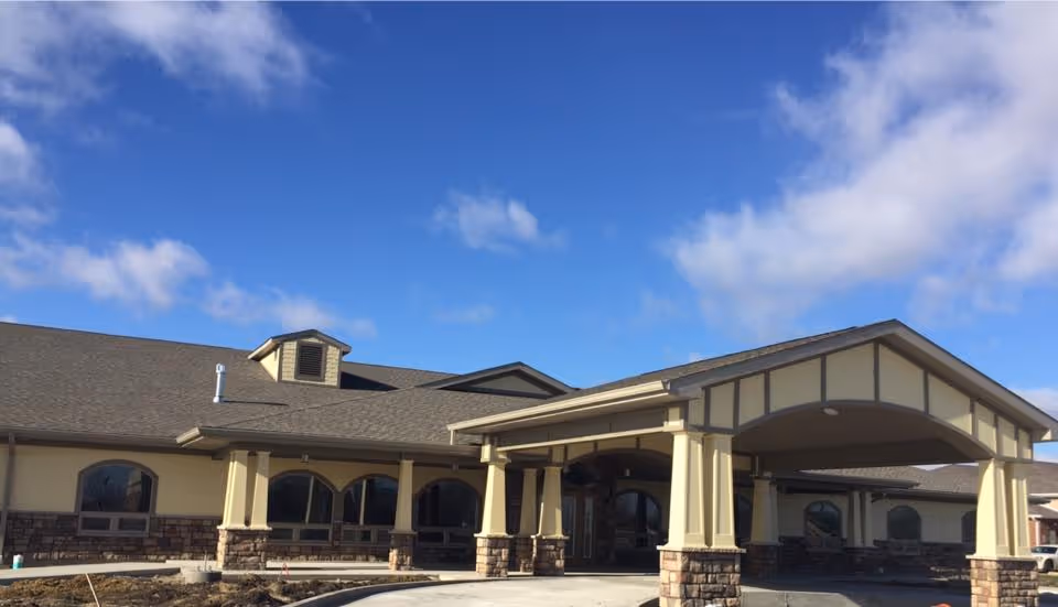 Single-story beige building with a covered porte-cochere supported by stone pillars under a blue sky.