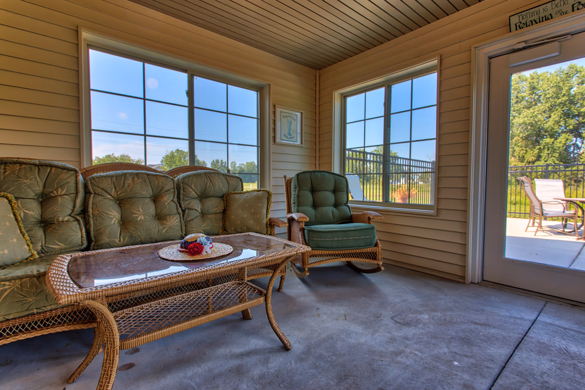 Sunroom with wicker sofa and chair, a glass-top coffee table, large windows and a door opening to an outdoor patio.