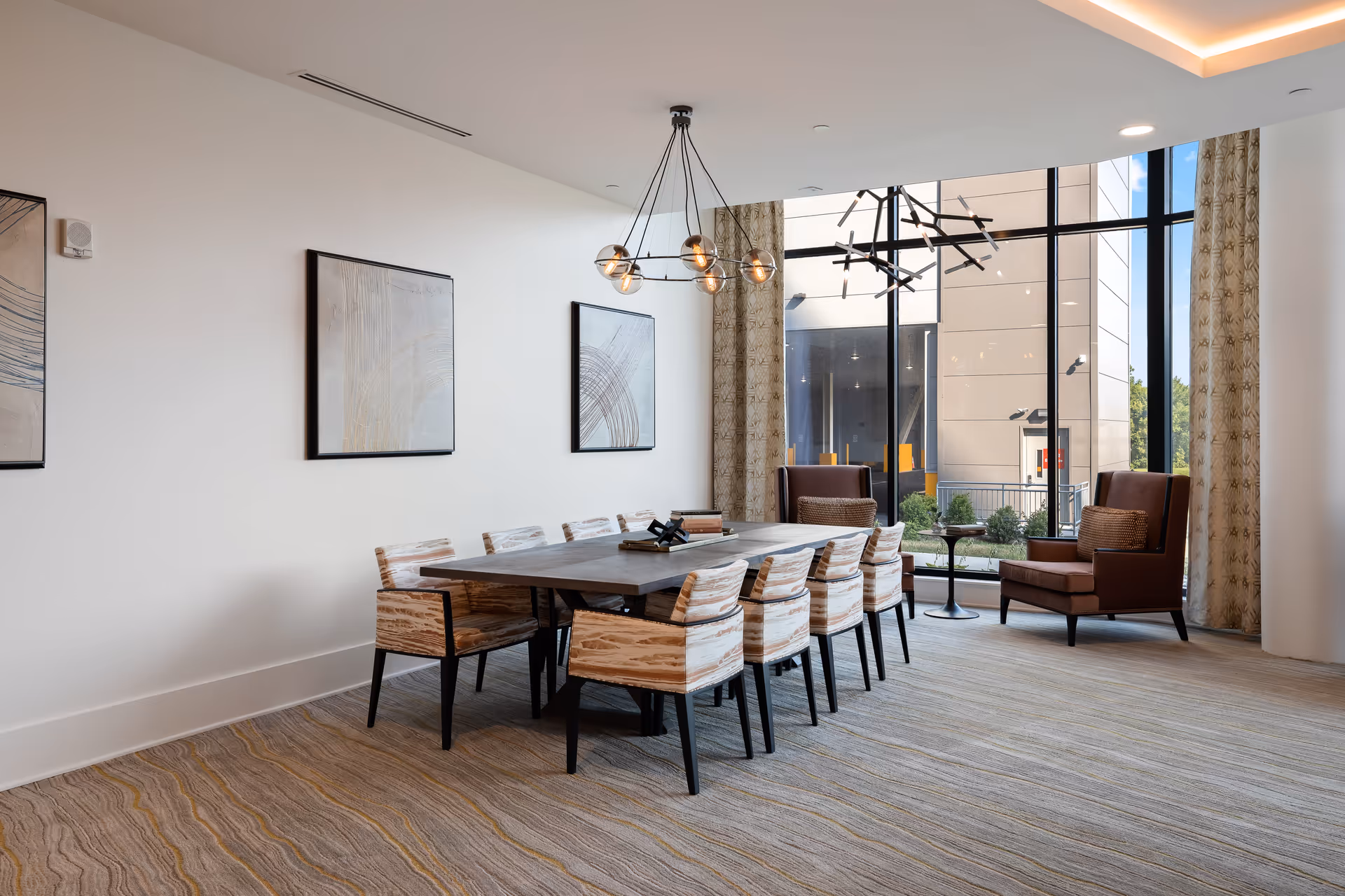 Bright communal dining room with a long table surrounded by chairs, pendant lighting, and large floor-to-ceiling windows.