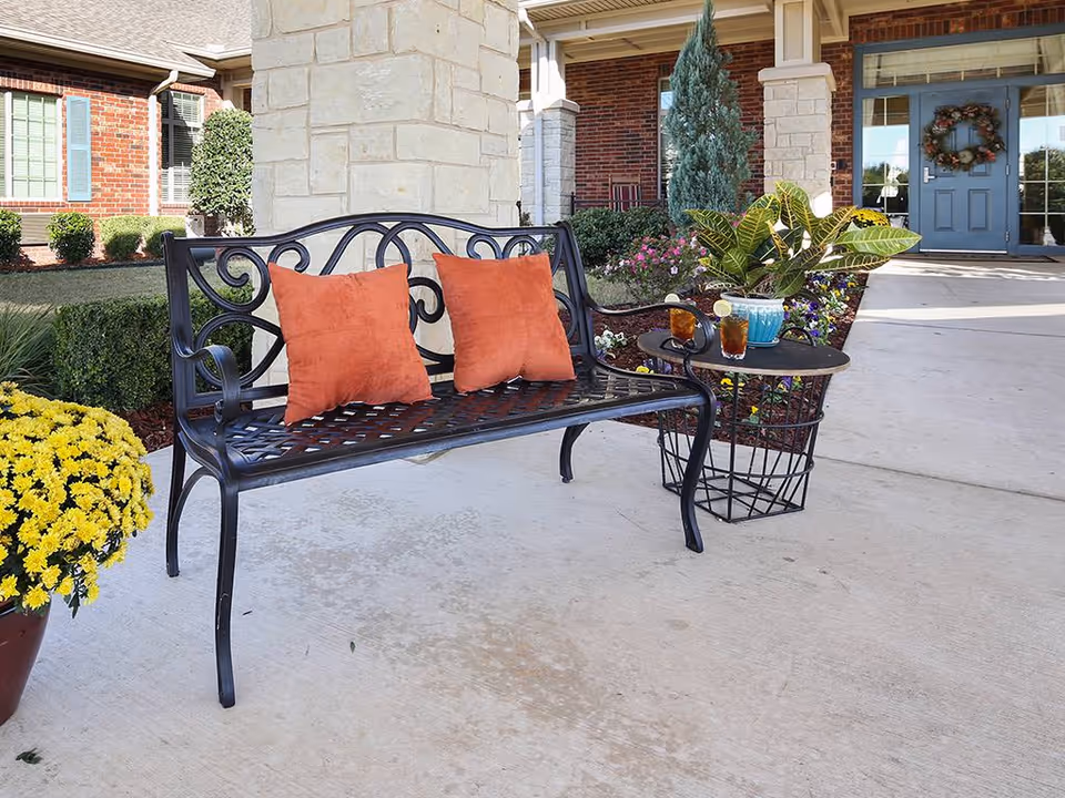 A metal bench with two orange pillows and a small side table holding drinks and a plant sits on the covered entryway of a brick building with a blue door.