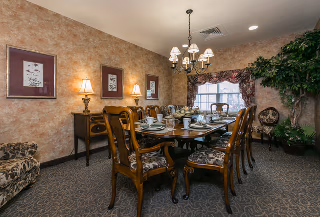 Elegantly furnished dining room with a long wooden table set for a meal, upholstered chairs, a chandelier, framed wall art, and a window with drapes.