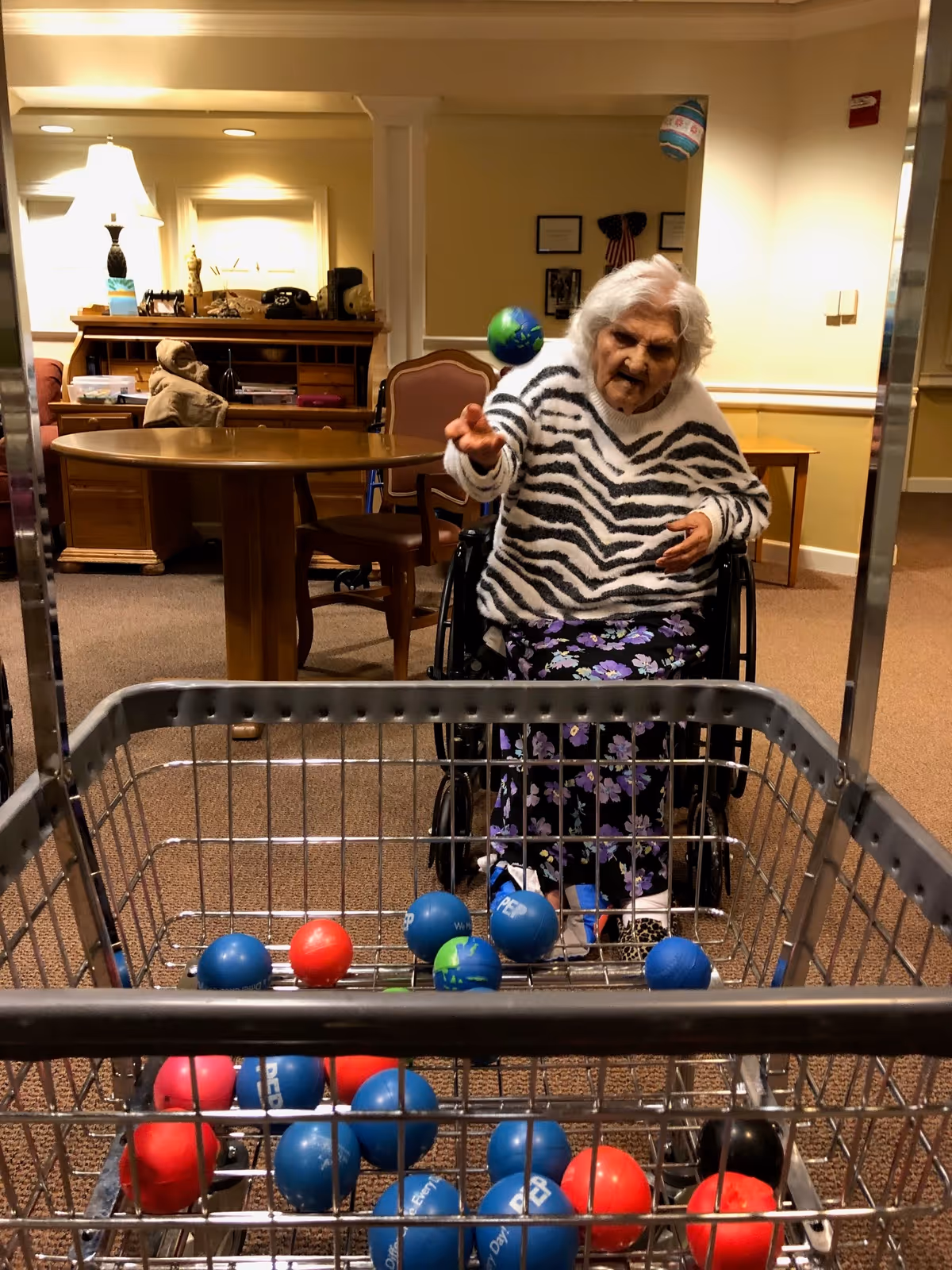 An elderly woman in a wheelchair wearing a zebra-striped sweater and floral pants is tossing a small ball towards a shopping cart filled with red and blue balls in a warmly lit room with wooden furniture and beige walls.