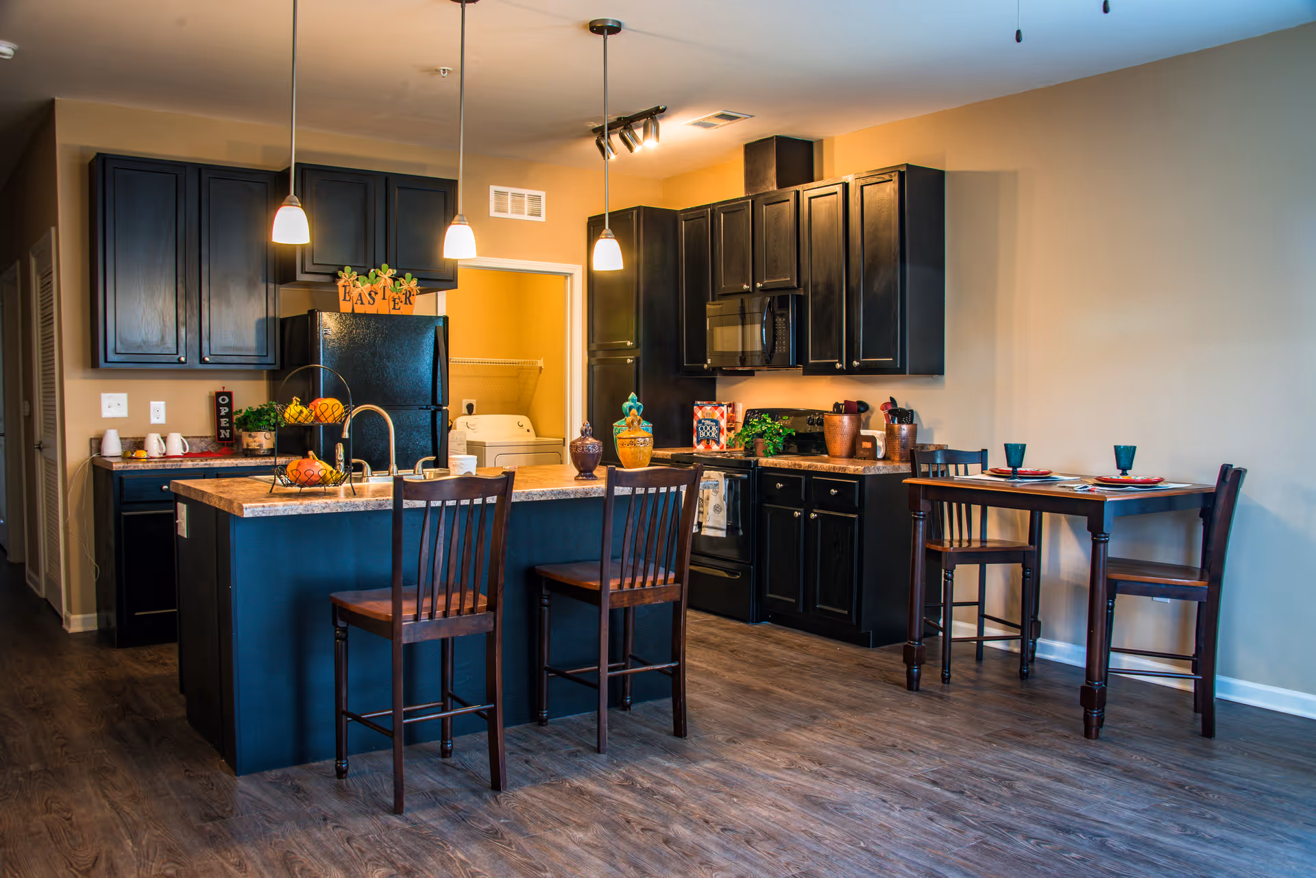 Open-plan kitchen with dark cabinets, a central island with bar stools and pendant lights, and a small dining table.