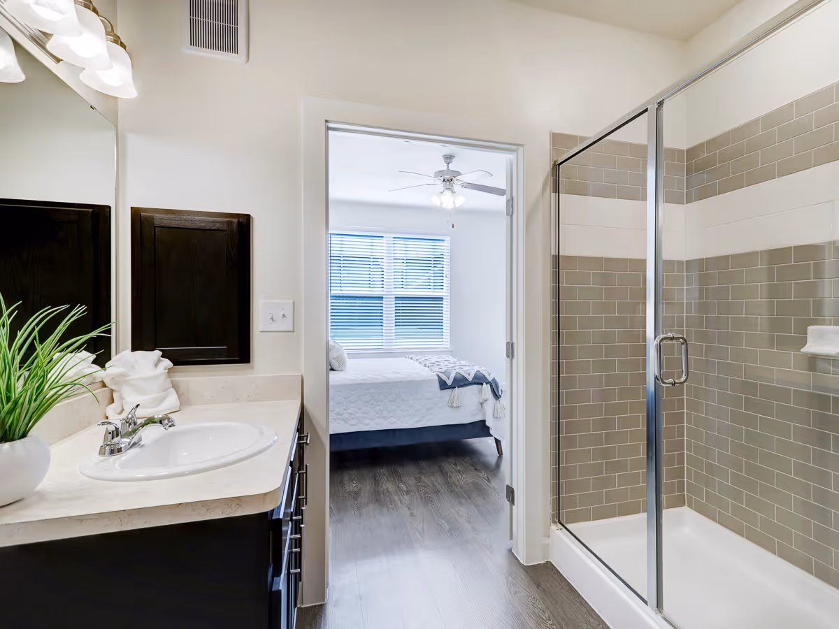 Bathroom with a sink and vanity on the left, a glass-enclosed tiled shower on the right, and a doorway showing a bedroom with a bed and window ahead.