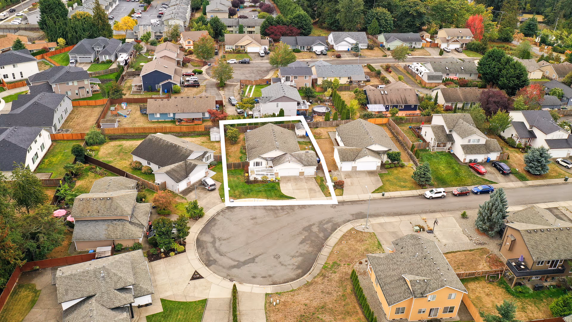 Aerial view of a residential neighborhood showing multiple houses with yards, driveways, and parked cars. One house is outlined in white, located at the end of a cul-de-sac. The area is surrounded by trees and greenery.