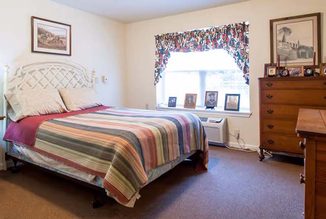 Bright, simply furnished bedroom with a bed covered by a striped quilt, a window with a valanced curtain and framed photos on the windowsill, and a wooden dresser.