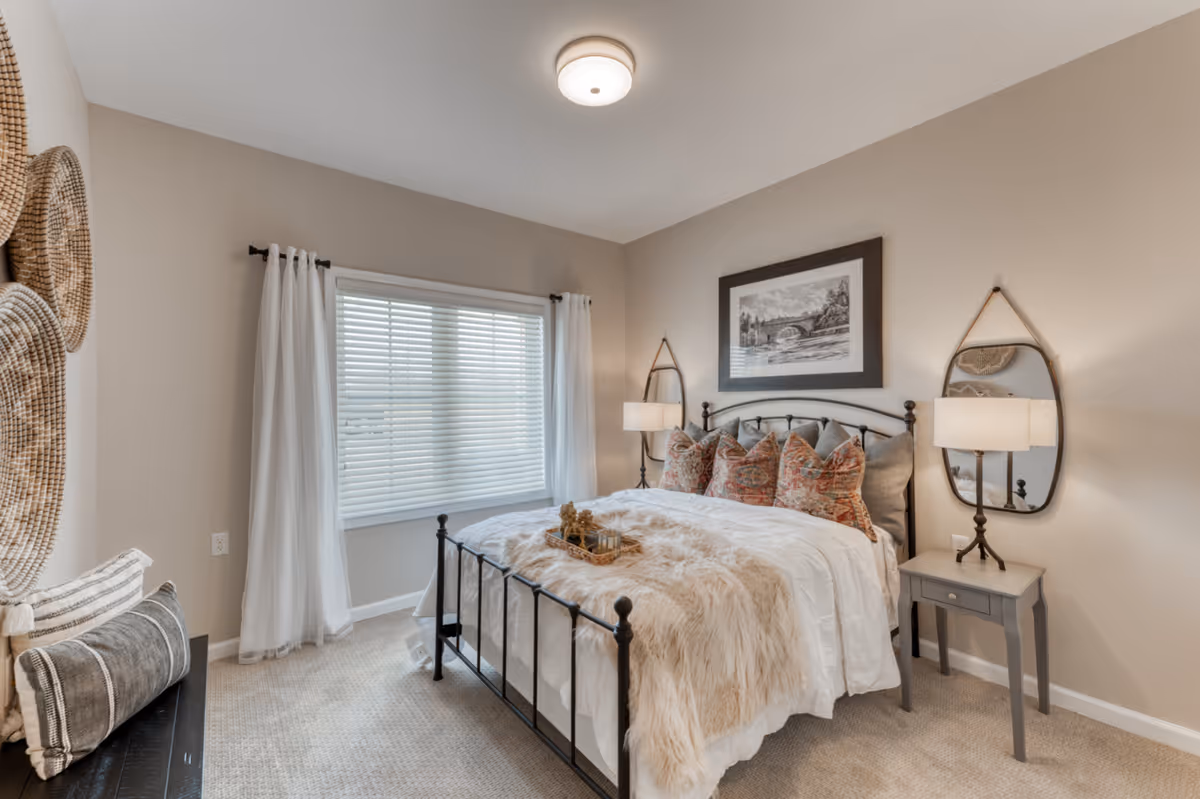 A cozy bedroom with a black metal bed frame, white bedding, and a beige faux fur throw. The bed is adorned with multiple decorative pillows. On either side of the bed are small gray nightstands with lamps. Above the bed hangs a framed black and white picture. Two oval mirrors hang on the wall behind the nightstands. A window with white blinds and sheer white curtains lets in natural light. The walls are painted beige, and the floor is carpeted. Decorative woven baskets hang on the left wall.