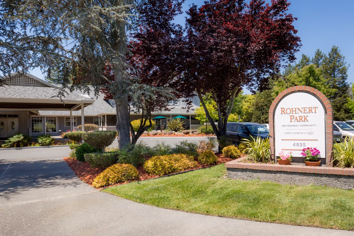 Exterior view of Cogir of Rohnert Park retirement community showing a landscaped entrance with trimmed bushes, trees, and a sign that reads 'Rohnert Park Retirement Community Happy Living by Cogir 4855'. The building and parking area are visible in the background under a clear blue sky.
