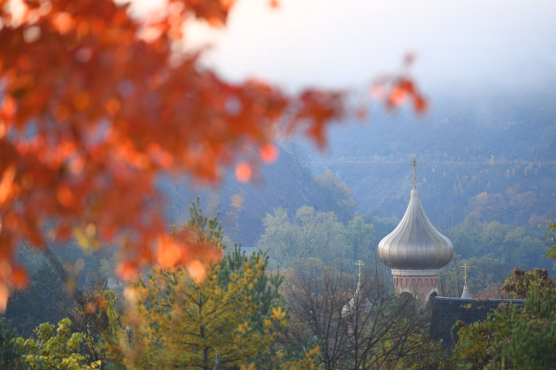 A scenic view of a silver onion dome church tower surrounded by trees with autumn foliage and a misty mountainous background.