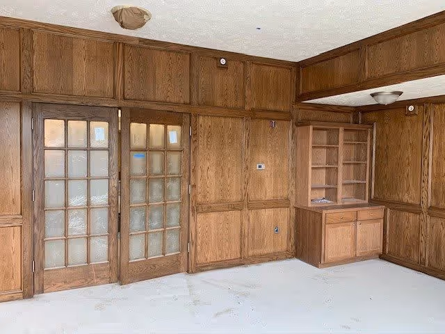 An empty room with wood-paneled walls and built-in wooden cabinets and shelves. There are two wooden framed glass doors on the left side. The floor is bare and appears unfinished, and the ceiling has two light fixtures.
