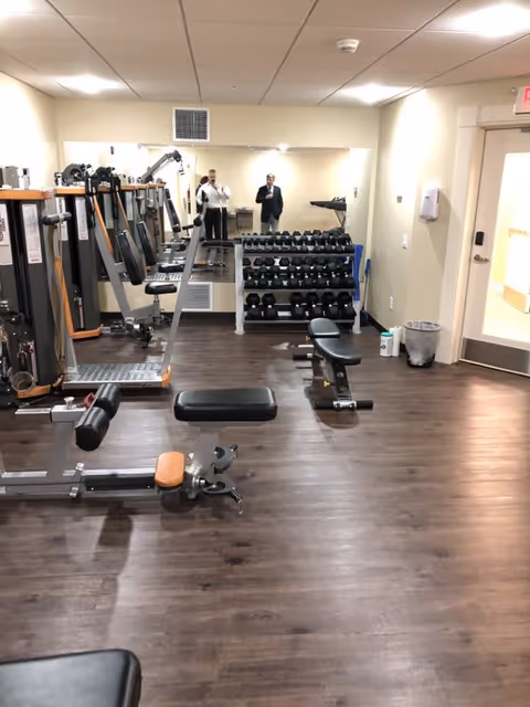 Interior fitness room with weight benches, strength machines, a dumbbell rack and a large mirror reflecting two people.