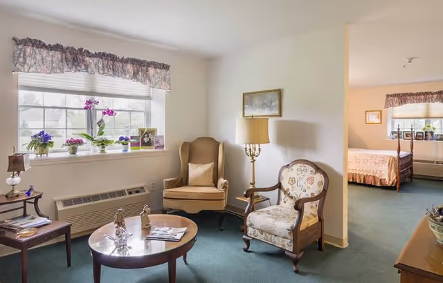 A cozy living area with two armchairs, a round wooden coffee table with decorative items, a side table with a lamp, and a large window with floral curtains and potted plants on the windowsill. In the background, a bedroom with a bed and a window with similar curtains is visible.