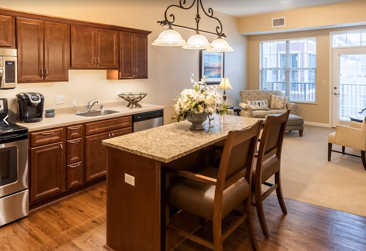 Interior view of a senior living facility showing a kitchen area with wooden cabinets, stainless steel appliances, a granite countertop island with two chairs, and a floral centerpiece. In the background, there is a living room area with a sofa, armchair, side table with a lamp, large windows, and a glass door leading outside.