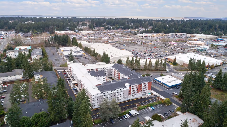 Aerial view of a multi-story residential building surrounded by parking lots, trees, and nearby commercial buildings.