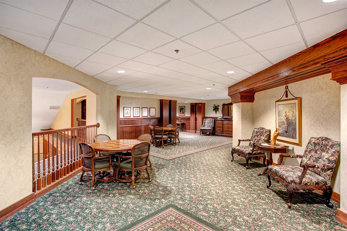 Interior view of a senior living facility lounge area with floral patterned carpet, wooden furniture including round tables with chairs, and upholstered armchairs. The walls are decorated with framed artwork and the ceiling has recessed lighting.