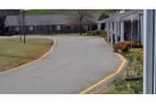 Curved driveway leading to a single-story building with a porch and columns, surrounded by grass and some shrubs.
