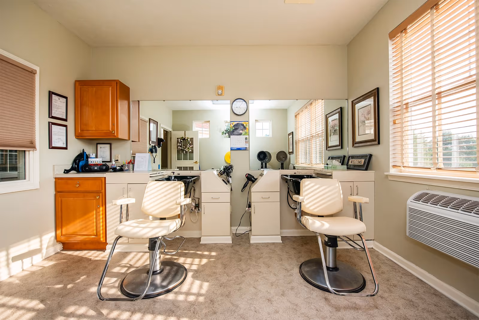 Interior view of a hair salon area in a senior living facility with two salon chairs facing large mirrors. The room has beige walls, carpeted floor, wooden cabinets, framed pictures on the walls, and windows with blinds letting in natural light. Hair dryers and salon equipment are visible on the counters.
