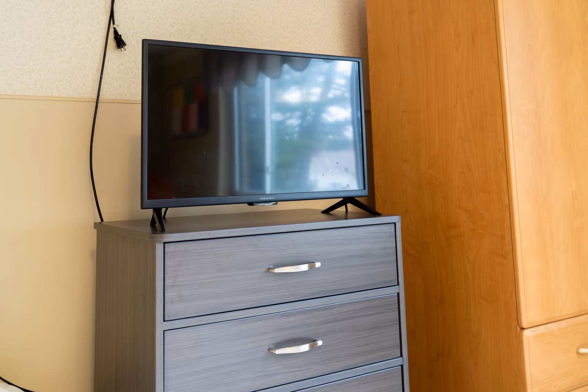 A flat-screen Insignia television sits on top of a gray wooden dresser with three drawers. To the right of the dresser is a wooden cabinet. The TV screen reflects a window with curtains and some outdoor scenery.