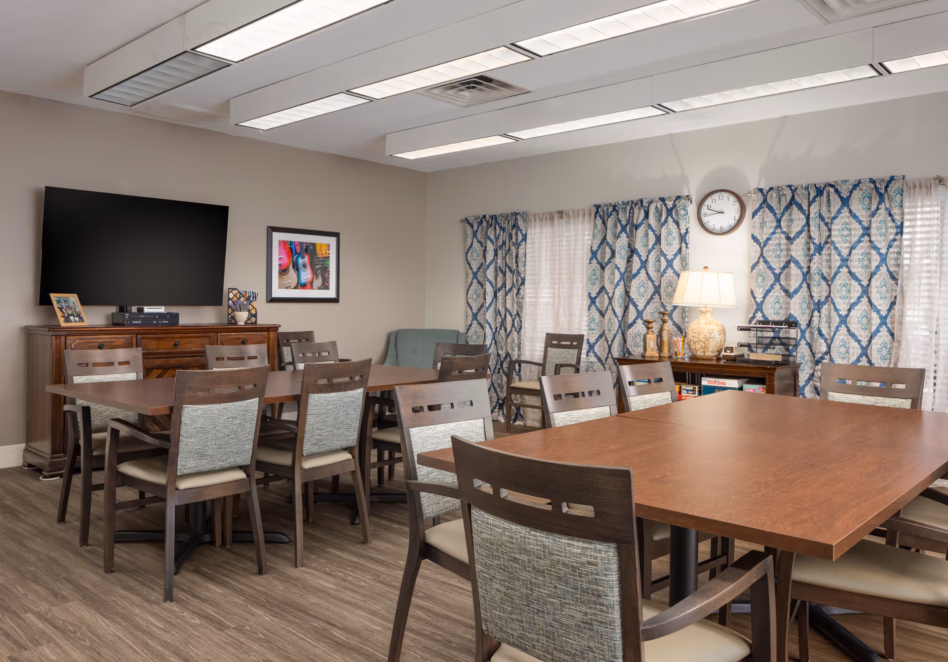 Community dining/activity room with multiple wooden tables and chairs, a wall-mounted TV, patterned curtains and a lamp on a sideboard.