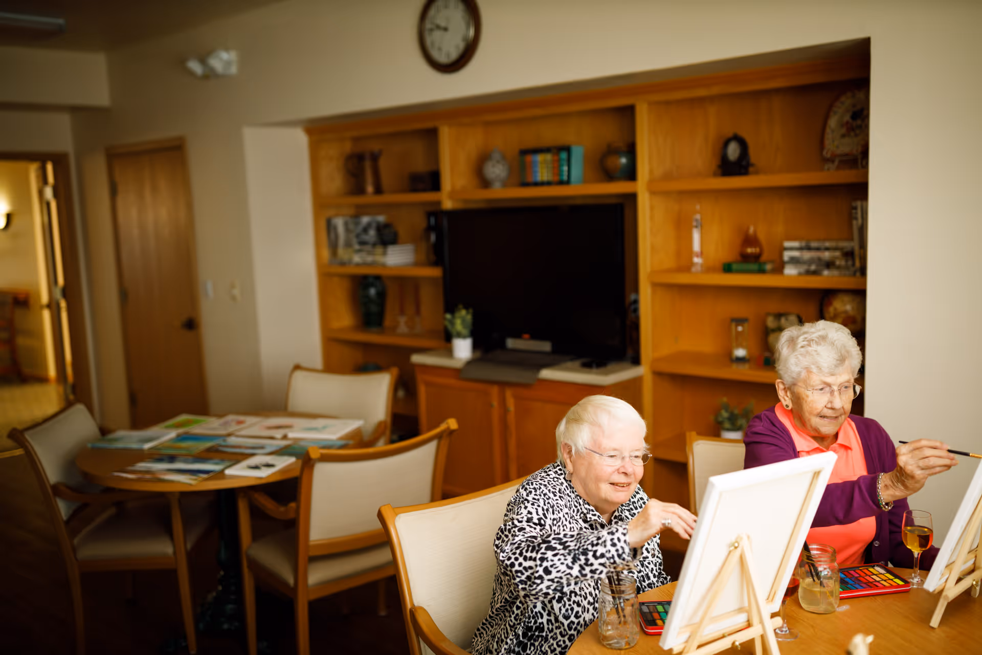 Two elderly women sitting at a table in a cozy room, painting on canvases with watercolors. Behind them is a wooden shelving unit with a TV, books, and decorative items. The room has a warm and inviting atmosphere.