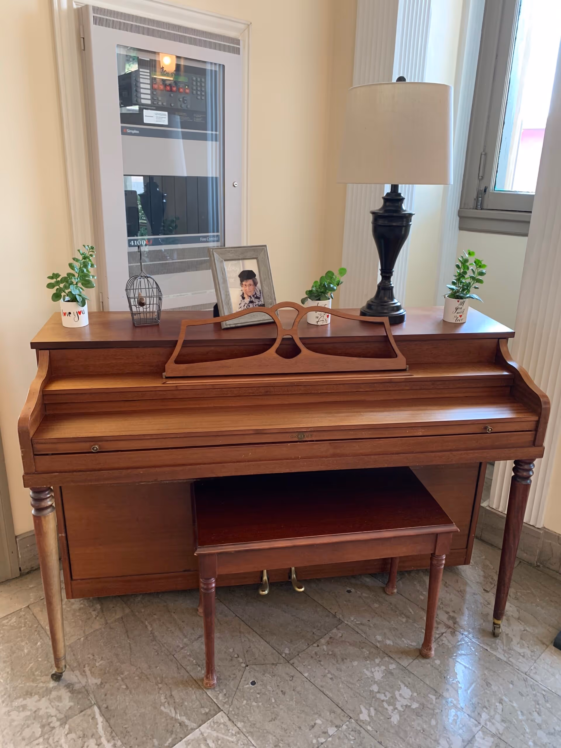 A wooden upright piano with a matching bench in front of it, placed on a tiled floor. On top of the piano are three small potted plants, a framed photo of an elderly woman, a small decorative birdcage, and a black table lamp with a white lampshade. Behind the piano is a fire control panel mounted on the wall and a window to the right.