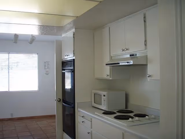 A kitchen area with white cabinets, a white countertop, a microwave, and a stovetop with four burners. There is a black built-in oven on the left side and a window with blinds in the adjacent room.
