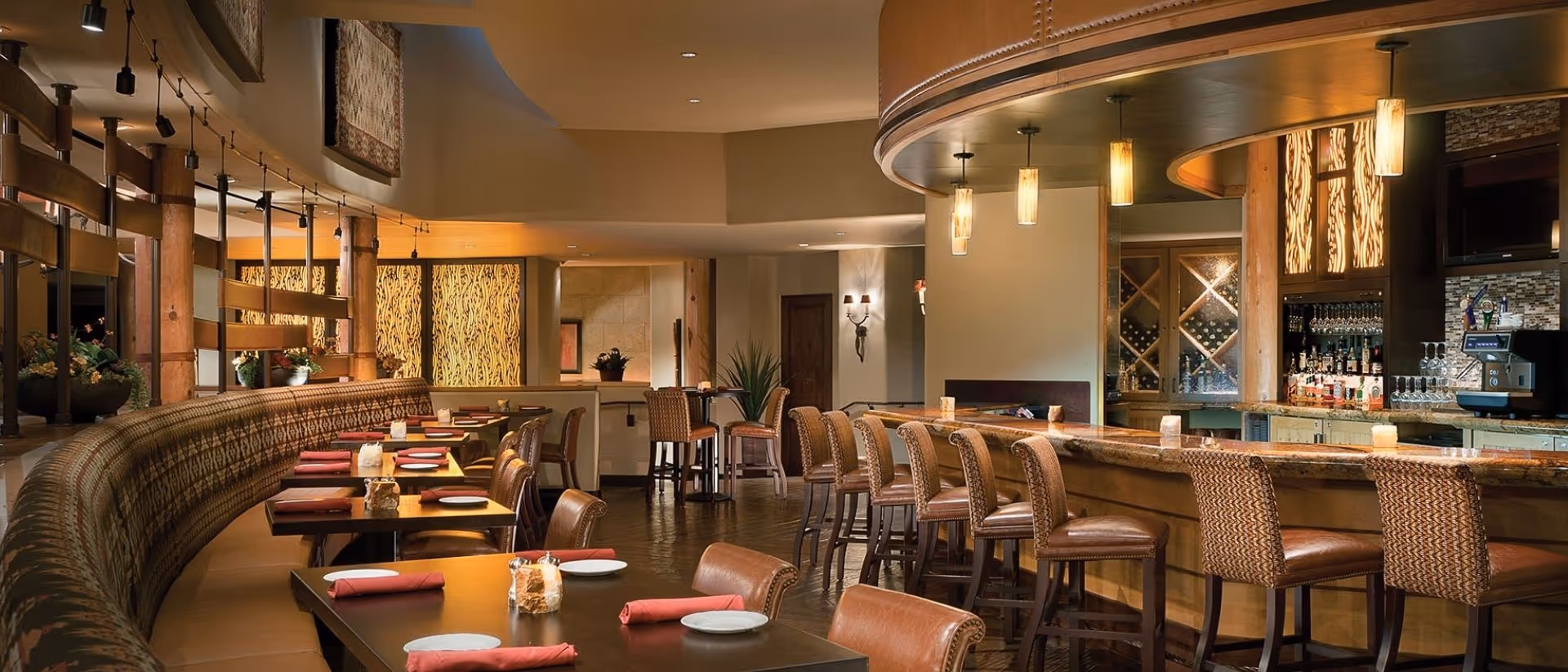 Interior view of a warm, inviting dining area with a curved booth seating along one side, tables set with plates and red napkins, and a bar with high chairs on the right side. The lighting is soft with hanging pendant lights above the bar and wall sconces, and there are decorative elements such as plants and textured wall panels.