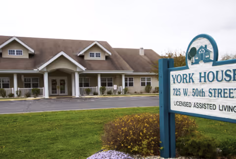 Exterior view of a single-story assisted living facility building with a covered entrance and multiple windows. In the foreground, there is a sign that reads 'YORK HOUSE 725 W. 50th STREET LICENSED ASSISTED LIVING' surrounded by grass and landscaping.