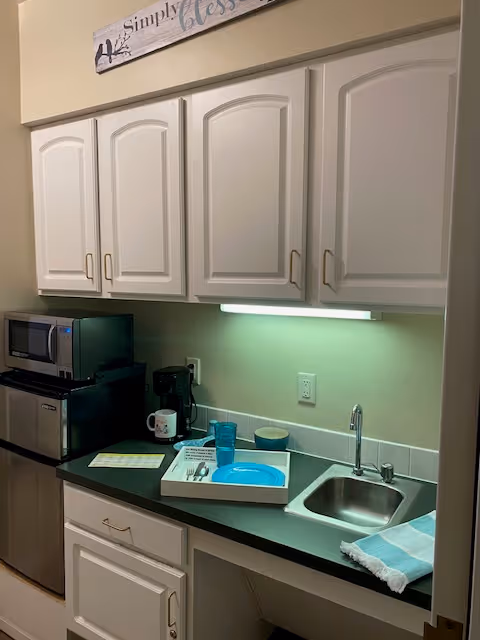 Small kitchenette area with white cabinets, a black countertop, a stainless steel sink with a faucet, a microwave, a mini refrigerator, a coffee maker, and a tray with a blue plate, cup, and utensils. A dish towel is placed next to the sink, and there is a wall-mounted light under the cabinets.