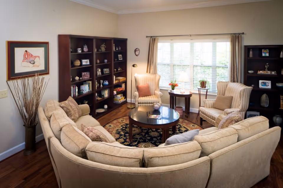 Cozy living room with a curved beige sectional sofa around a round coffee table, bookshelves, and two armchairs by a large window.