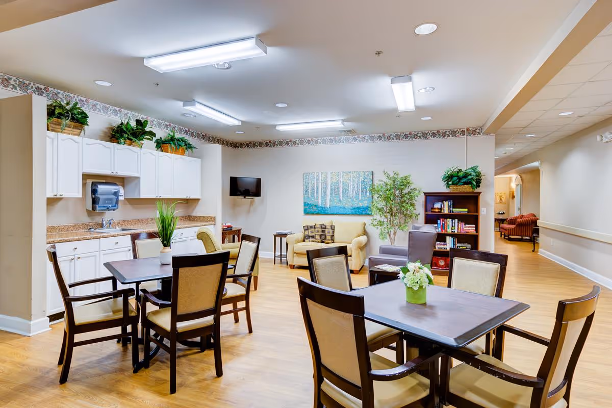 Bright communal dining and lounge area with tables and chairs, a small kitchenette, and seating near a TV and bookshelf.