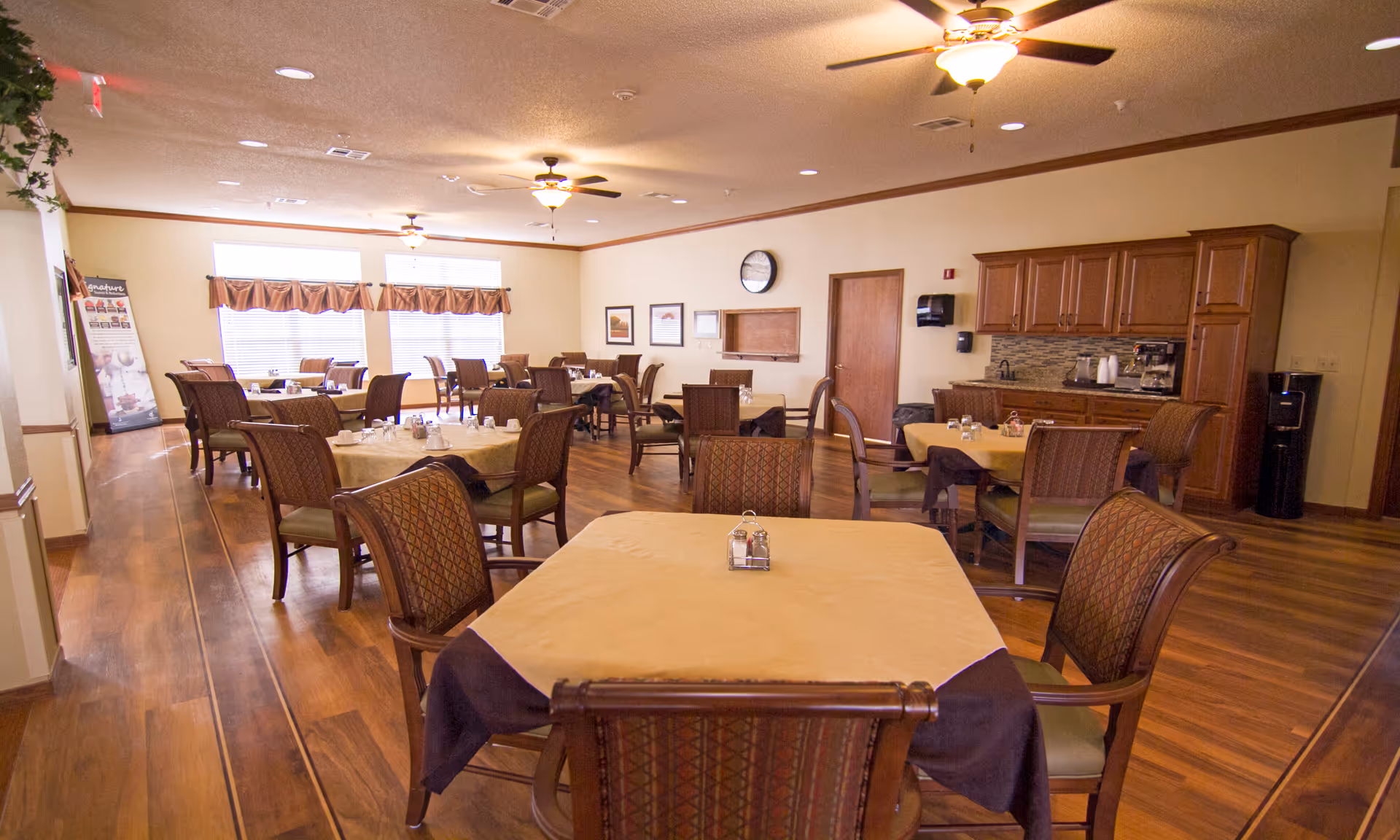 A spacious dining room with multiple tables covered with beige and dark brown tablecloths, surrounded by cushioned chairs. The room has wooden flooring, ceiling fans with lights, large windows with valances, and a kitchenette area with wooden cabinets and a coffee machine.