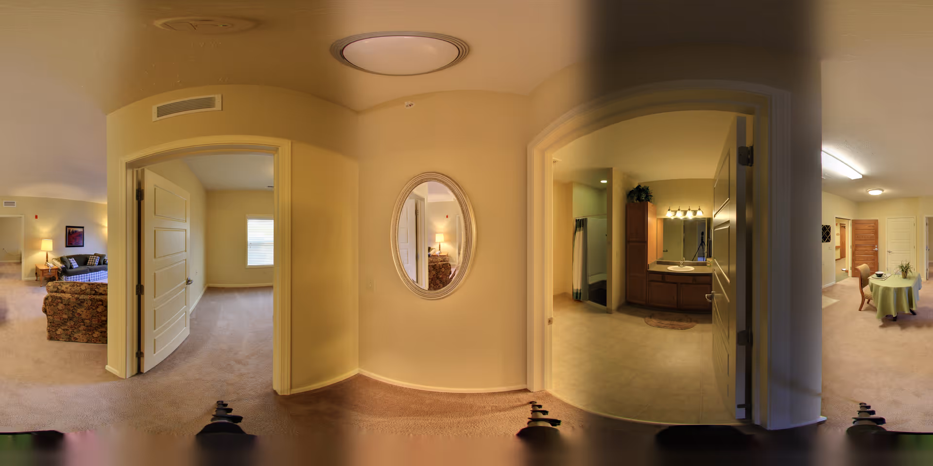 Panoramic interior hallway of a senior apartment showing doorways to a bedroom, a bathroom with a vanity, and a living/dining area with an oval wall mirror.