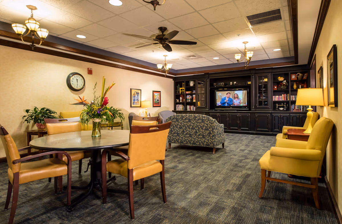 A cozy common area in a senior living facility featuring a round table with four tan chairs and a vase of flowers on top. The room has carpeted flooring, yellow armchairs along the right wall, and a patterned loveseat facing a large dark wood entertainment center with a TV displaying a man and woman. The walls are decorated with framed pictures and a clock, and the ceiling has recessed lighting, ceiling fans, and hanging light fixtures.