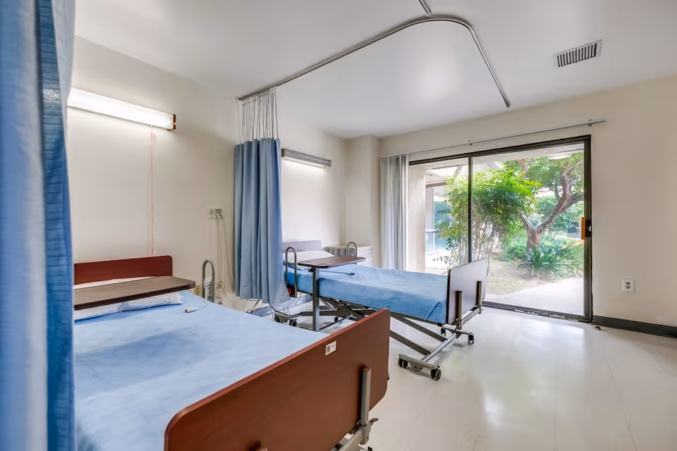 A hospital-style room with two medical beds covered in blue sheets, separated by blue privacy curtains. The room has a large sliding glass door that opens to a garden area with trees and greenery outside. The walls are light-colored, and the floor is tiled.