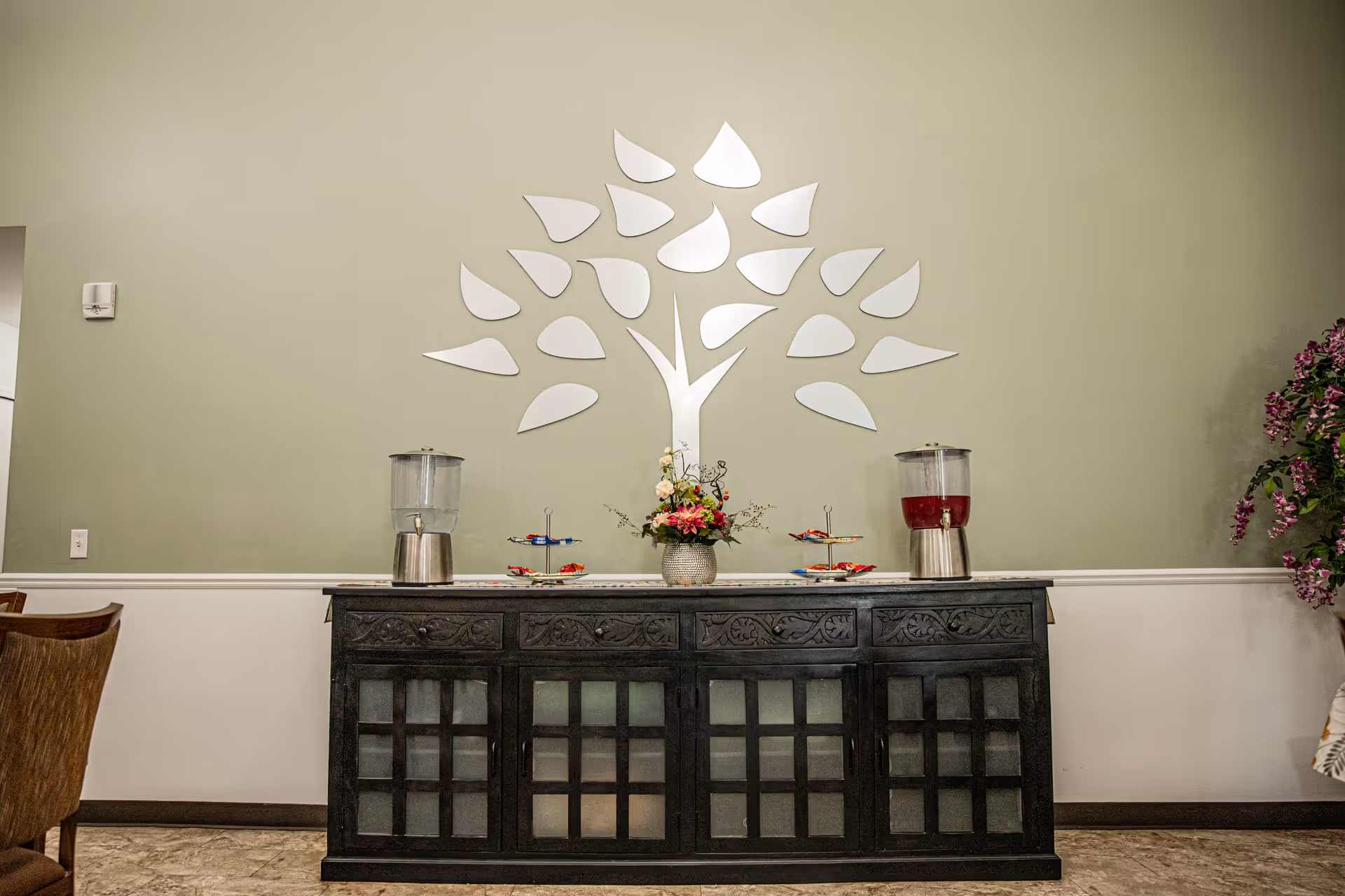 A black carved sideboard topped with beverage dispensers and a floral arrangement sits against a pale green wall decorated with a stylized mirrored tree.